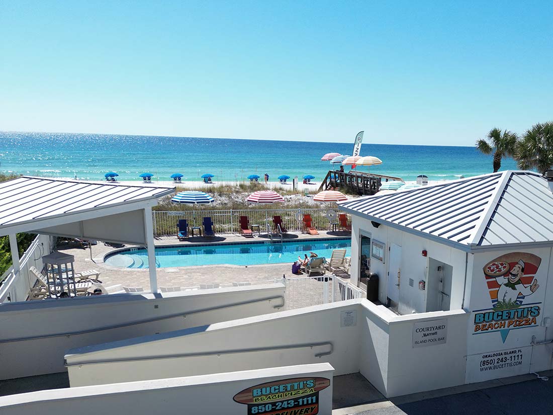 Beachside view of a pool, beach umbrellas, and the ocean on a sunny day with clear blue skies.