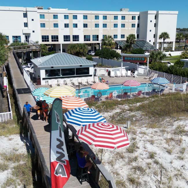A beachside hotel pool with colorful umbrellas, a pizza flag, and people on the wooden boardwalk.