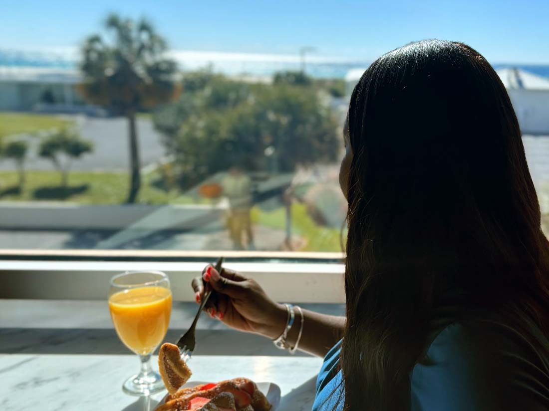 Woman enjoying breakfast by the window at Sundrift Inn with a sunny beachside view.